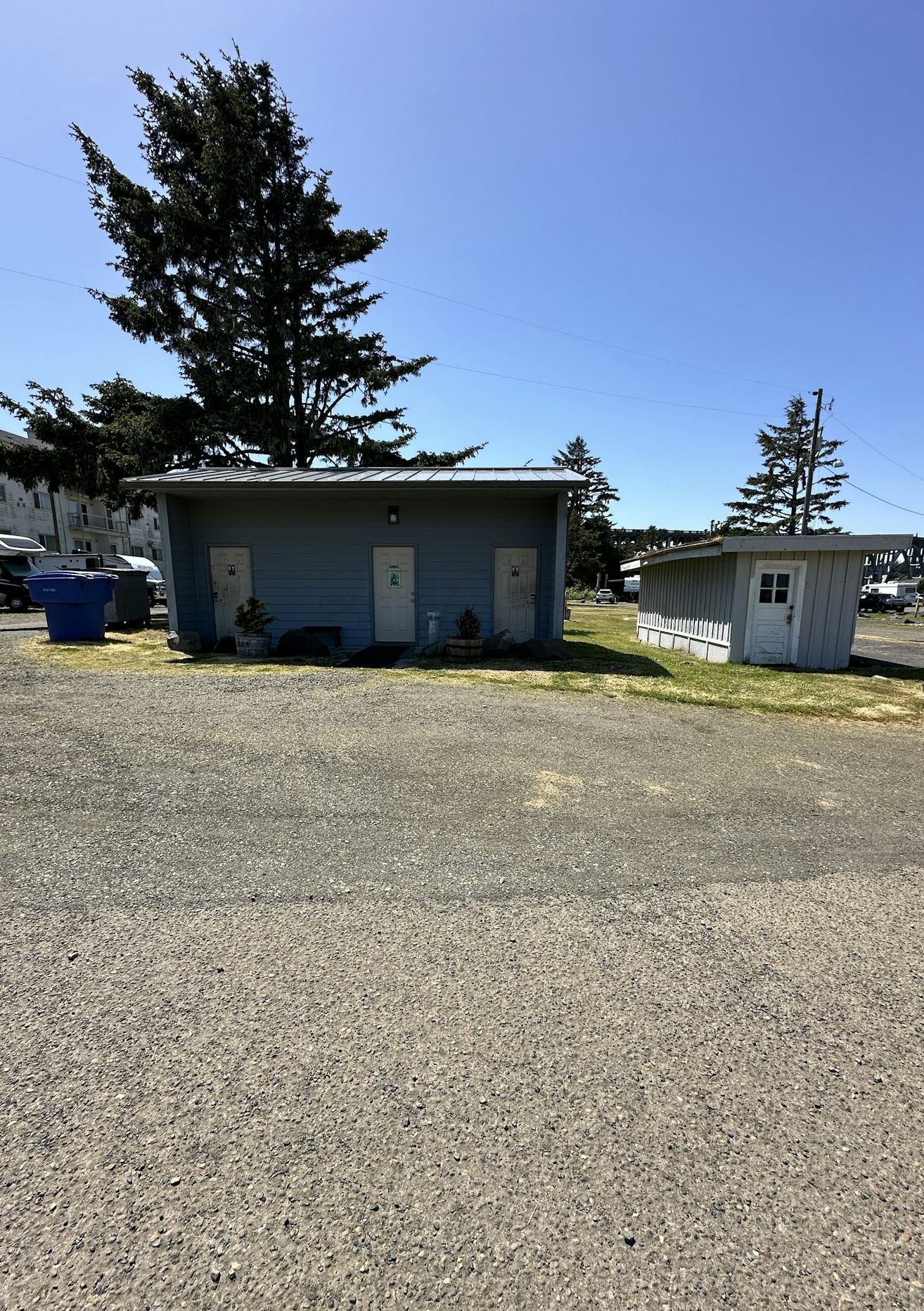 A small blue building with two doors, a large tree behind it, a gravel lot, and a white shed on a sunny day.