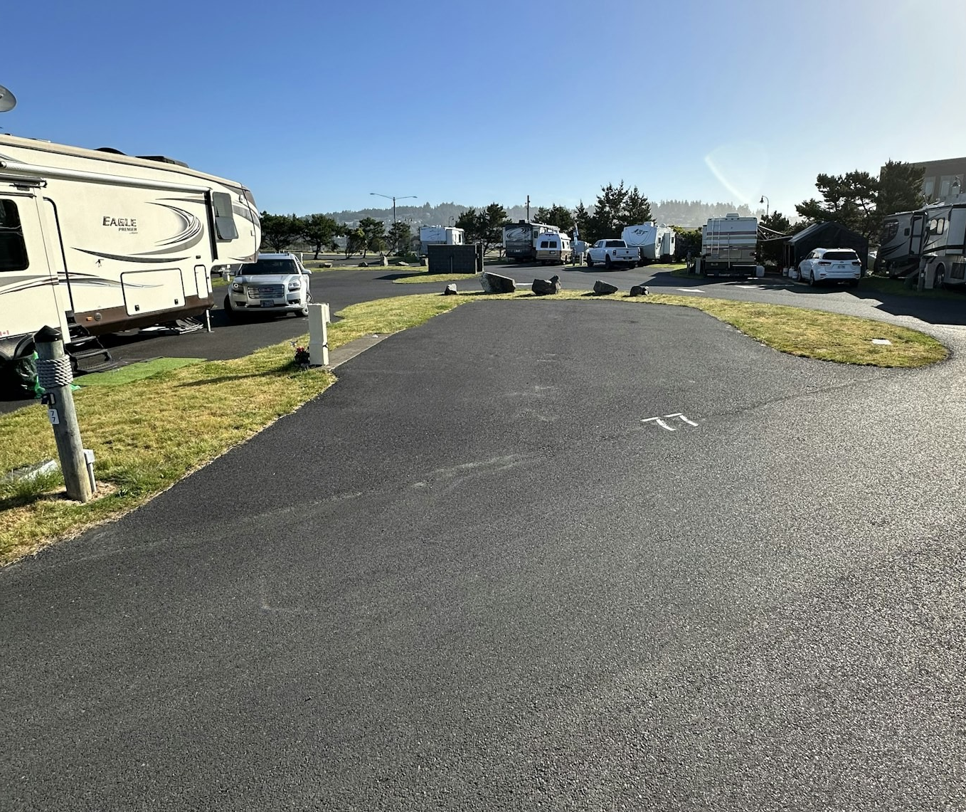 Sunny RV park with multiple campers and vehicles, green grass patches, asphalt roads, and utility posts.
