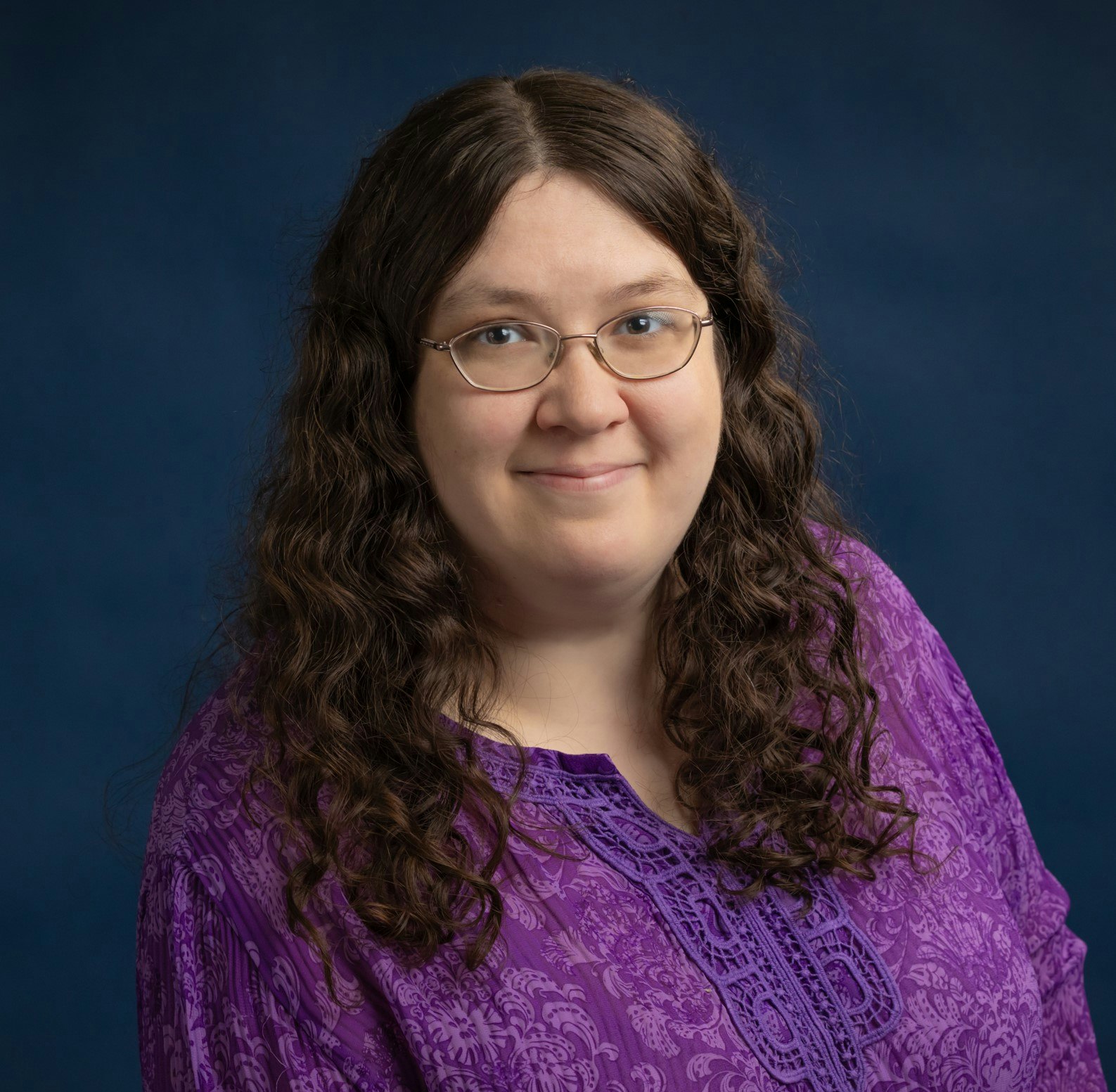 A woman smiling, wearing glasses and a purple blouse, with a blue background.