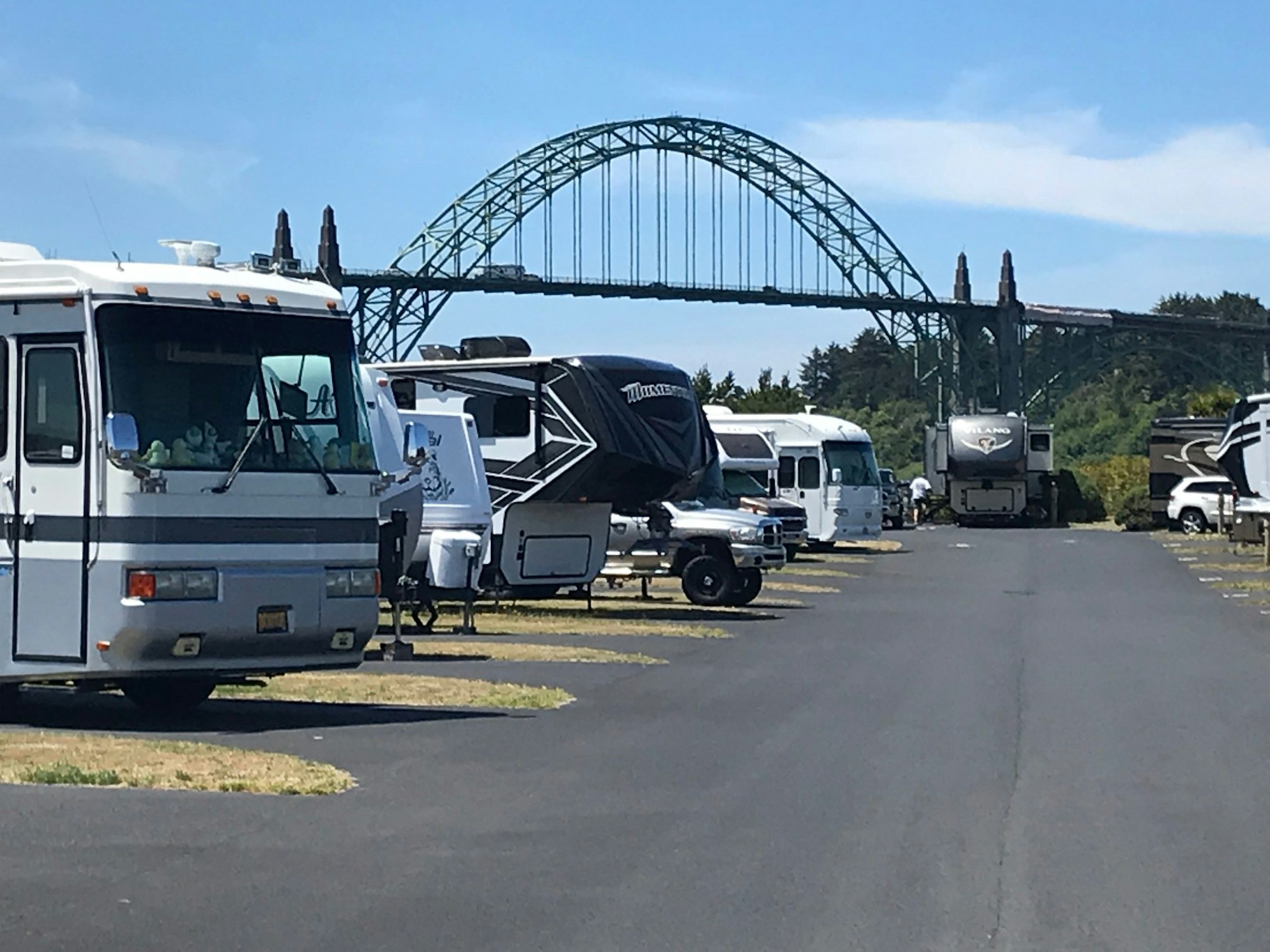 A row of RVs parked along a road with a green bridge in the background under a clear sky.