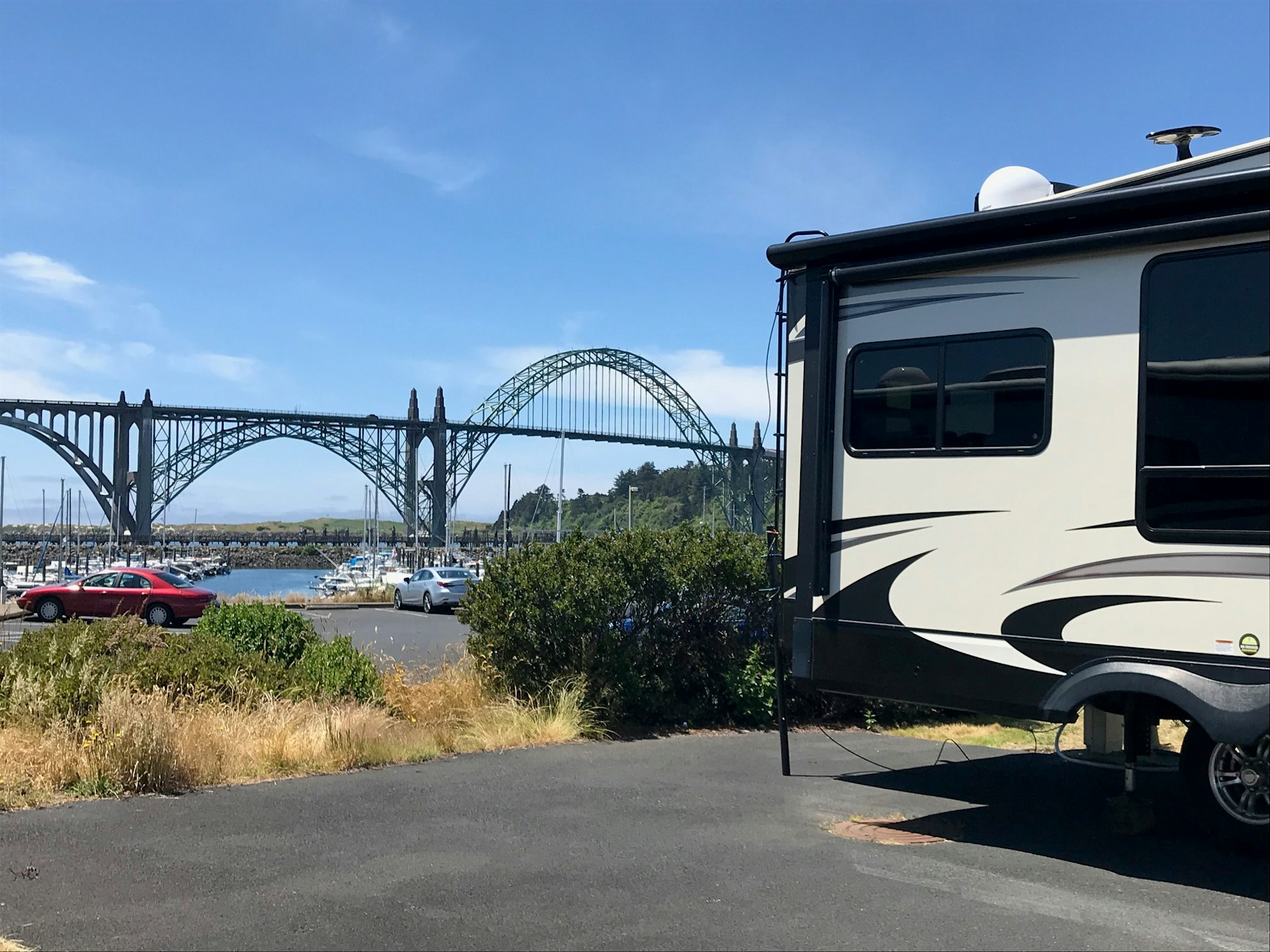 An RV parked near a marina with boats, set against a bridge and clear blue sky.