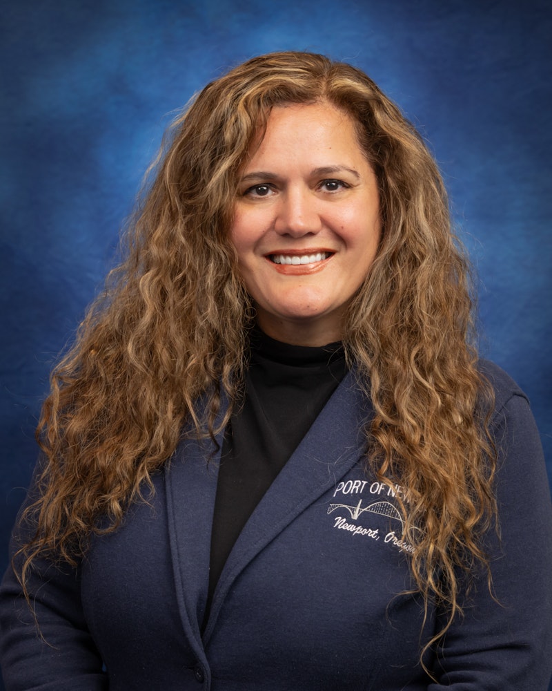 A person with curly hair, smiling, wearing a blue jacket with "Port of Newport" logo on a blue background.
