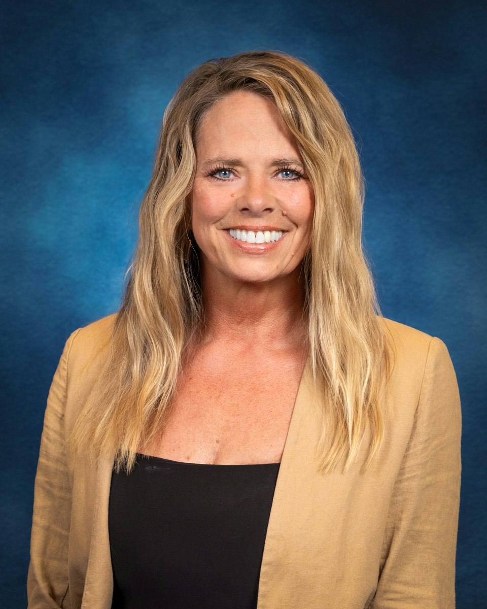 A woman with wavy hair smiles in front of a blue backdrop, wearing a beige blazer over a black top.