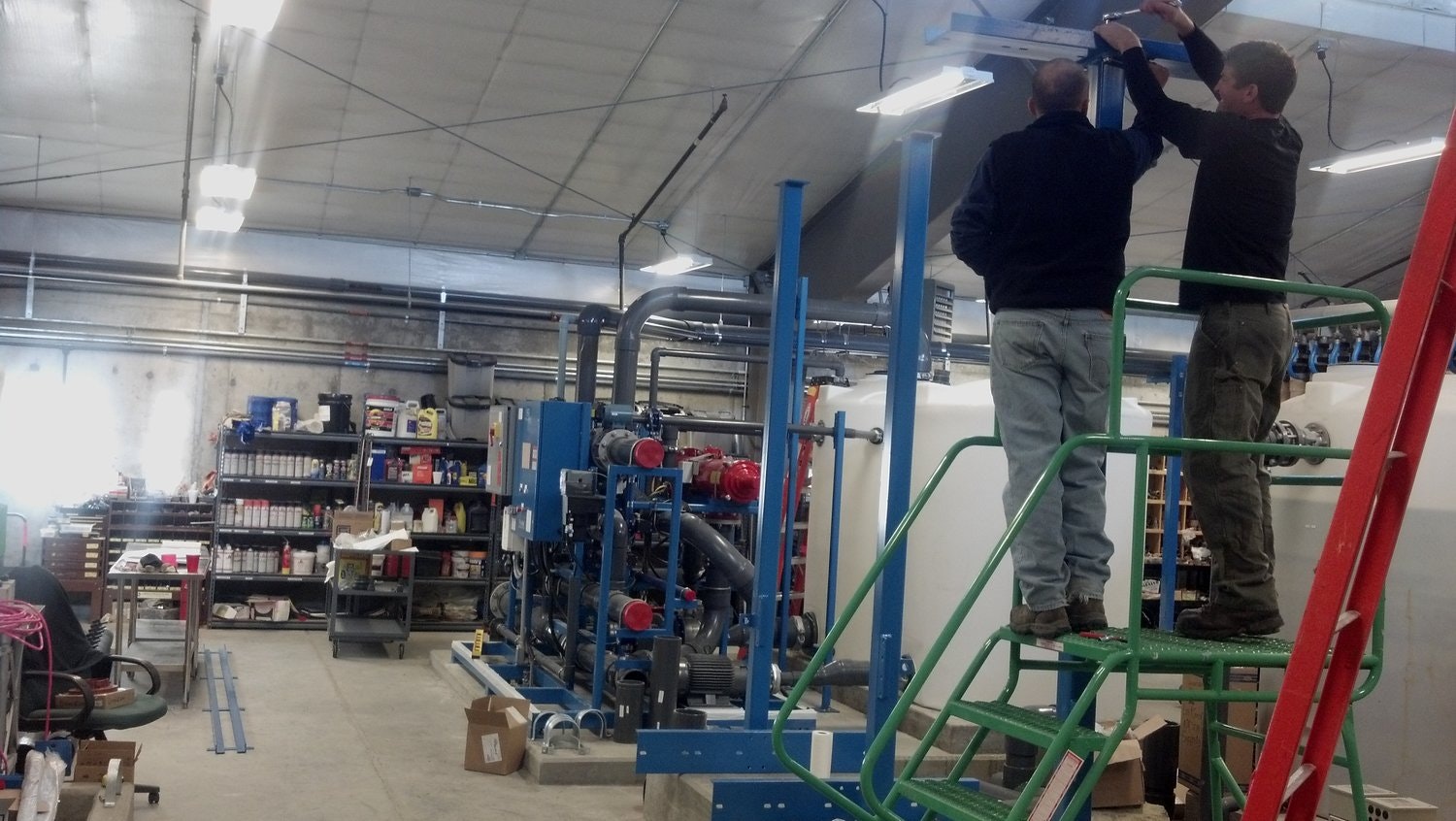 Two individuals are installing equipment in a warehouse filled with tools and machinery, with shelves of supplies in the background.