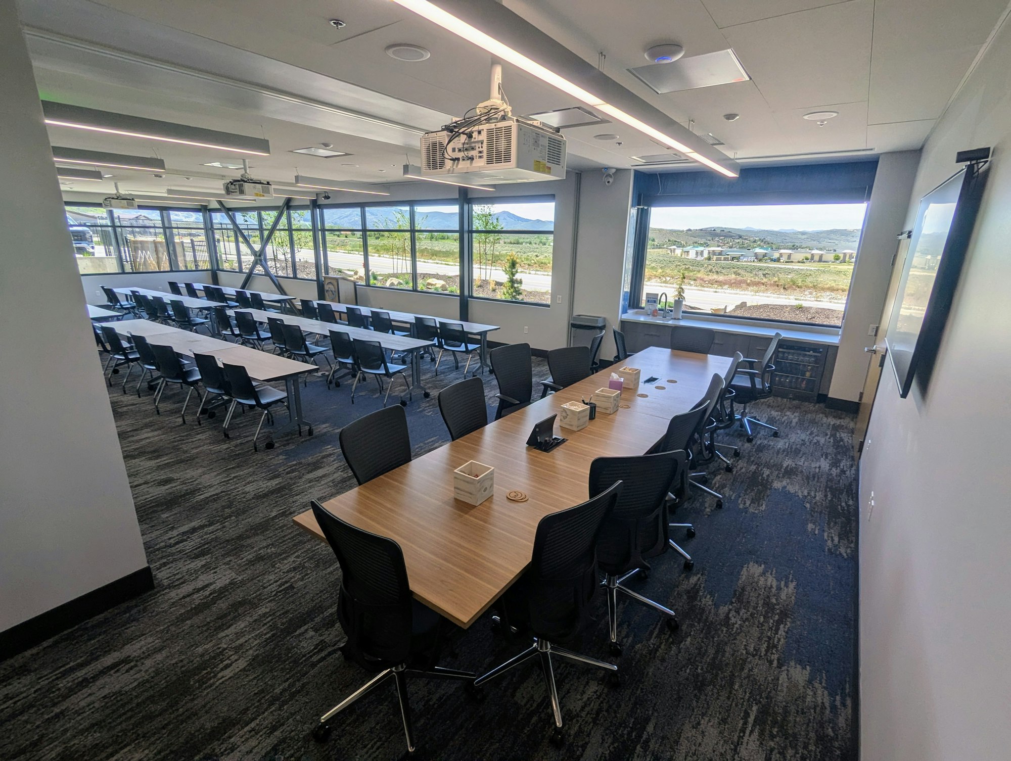 A modern meeting room with tables, chairs, a projector, and windows offering views of greenery and mountains outside.