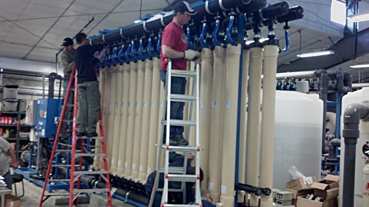Workers are installing equipment, using ladders, in an industrial setting with large pipes and machinery in the background.
