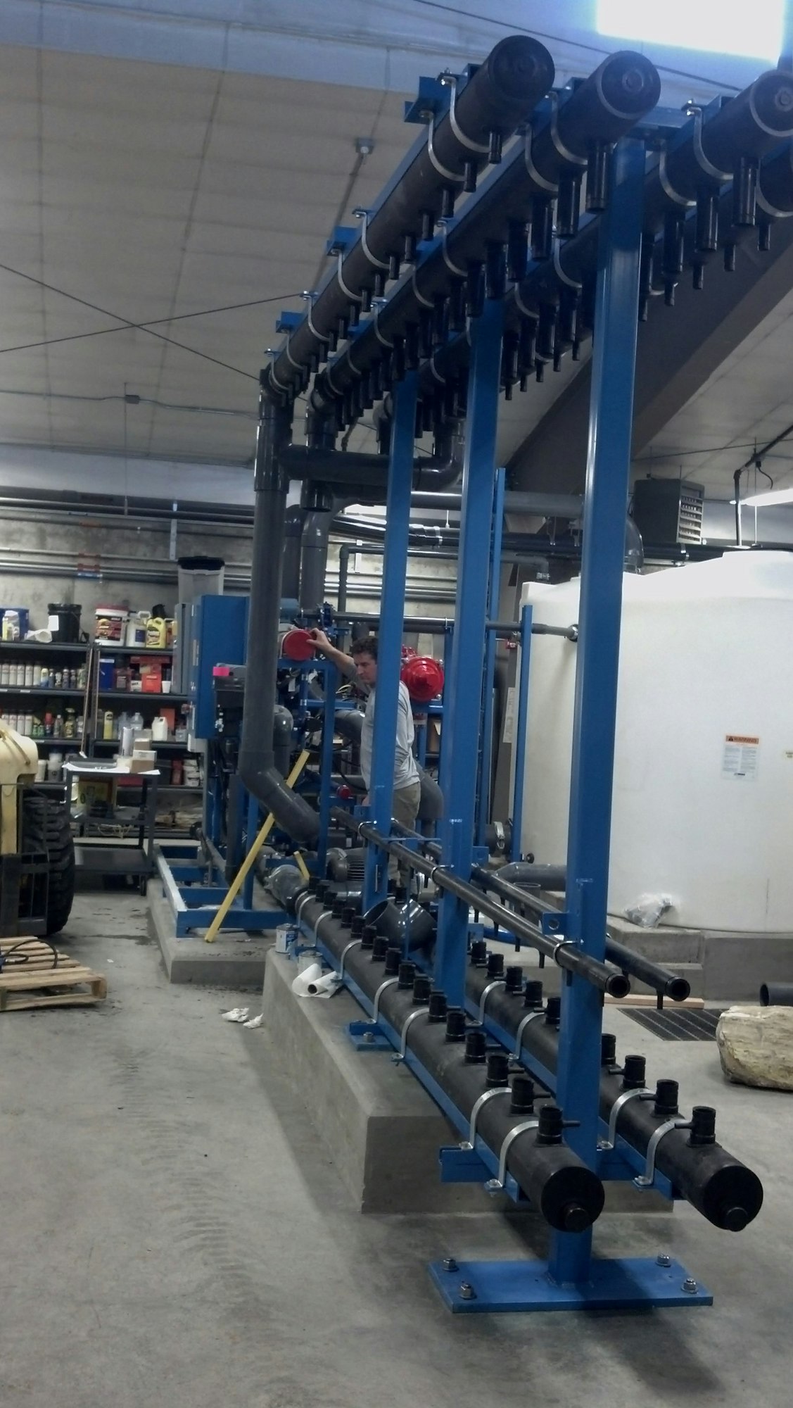 The image shows a mechanical setup with pipes, valves, and a person working. It appears to be part of a workshop or industrial area.