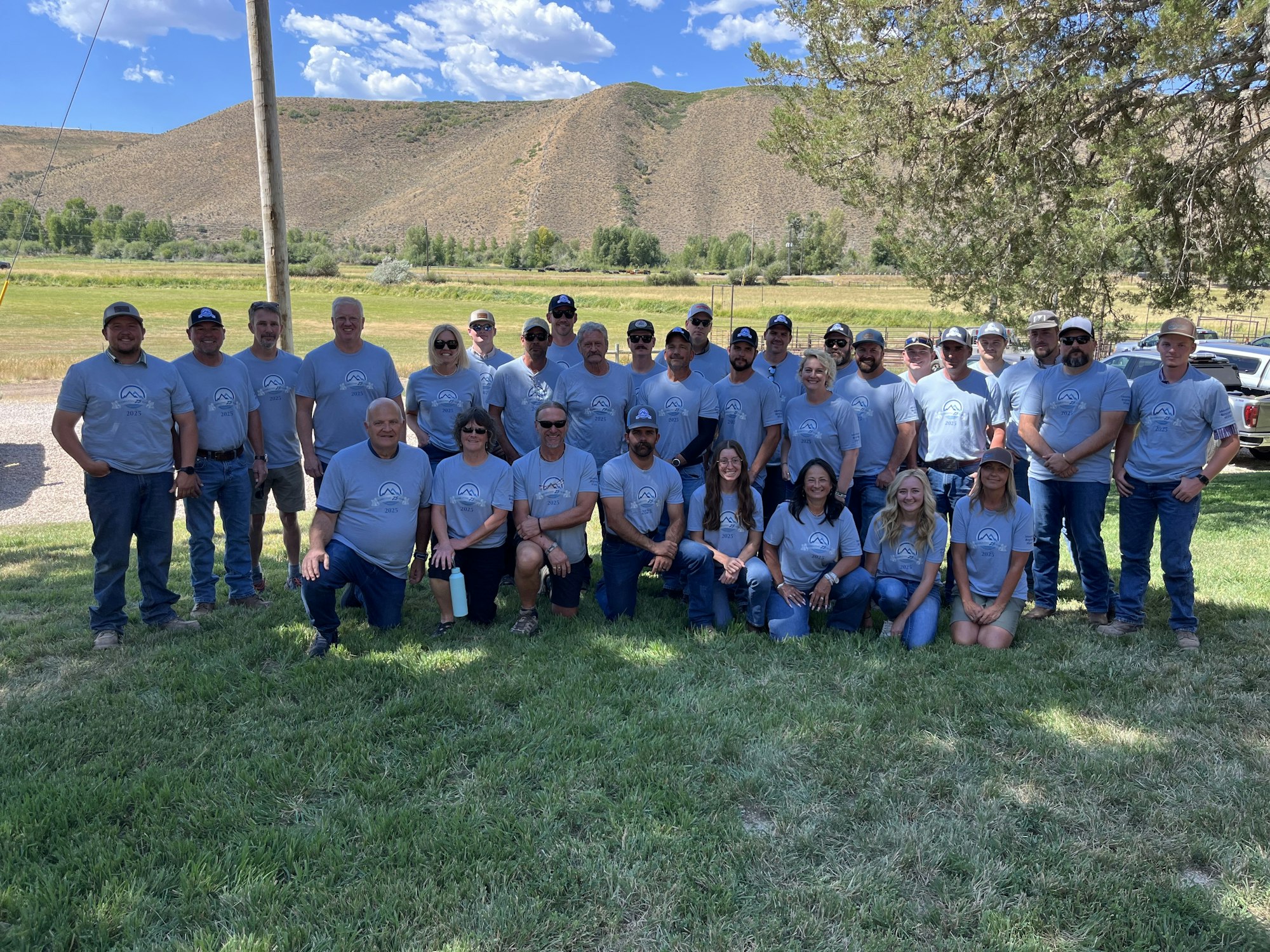 A group of people in matching light blue shirts posing outdoors on grass, with hills and trees in the background.