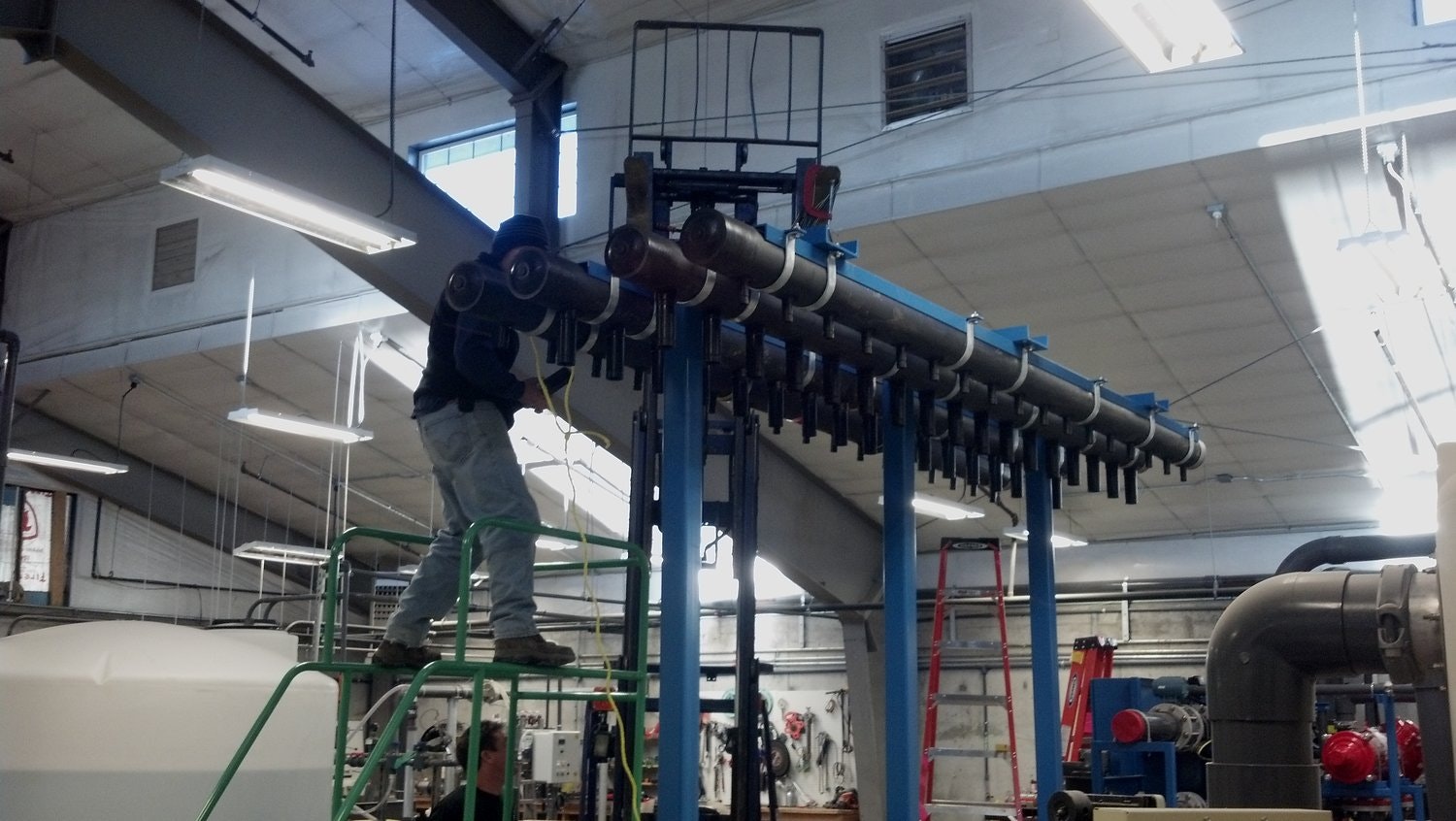 The image shows a person working on a metal assembly using a ladder and machinery in a factory setting.