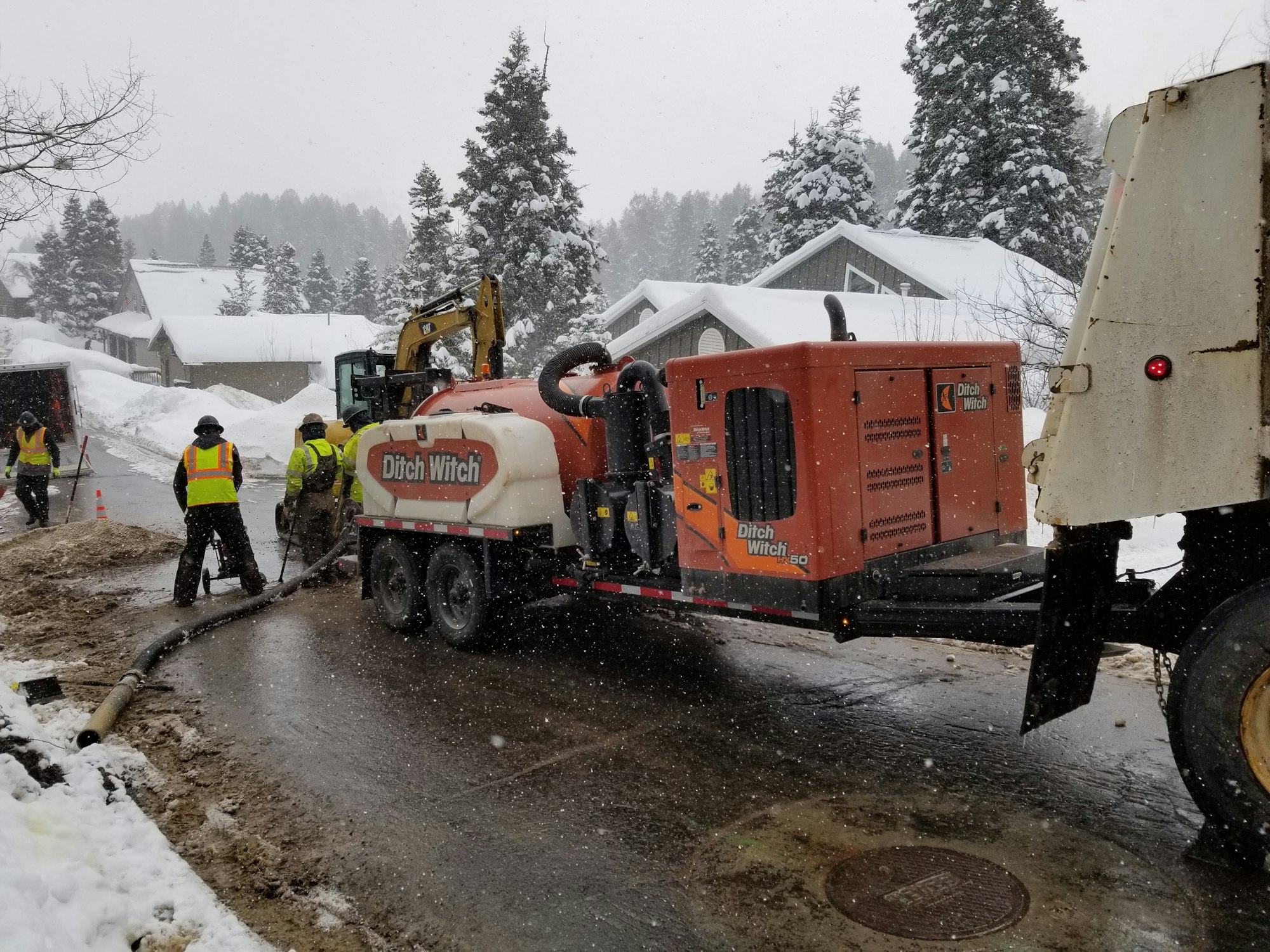 Workers using construction equipment, including a "Ditch Witch," during snowy weather on a residential street.
