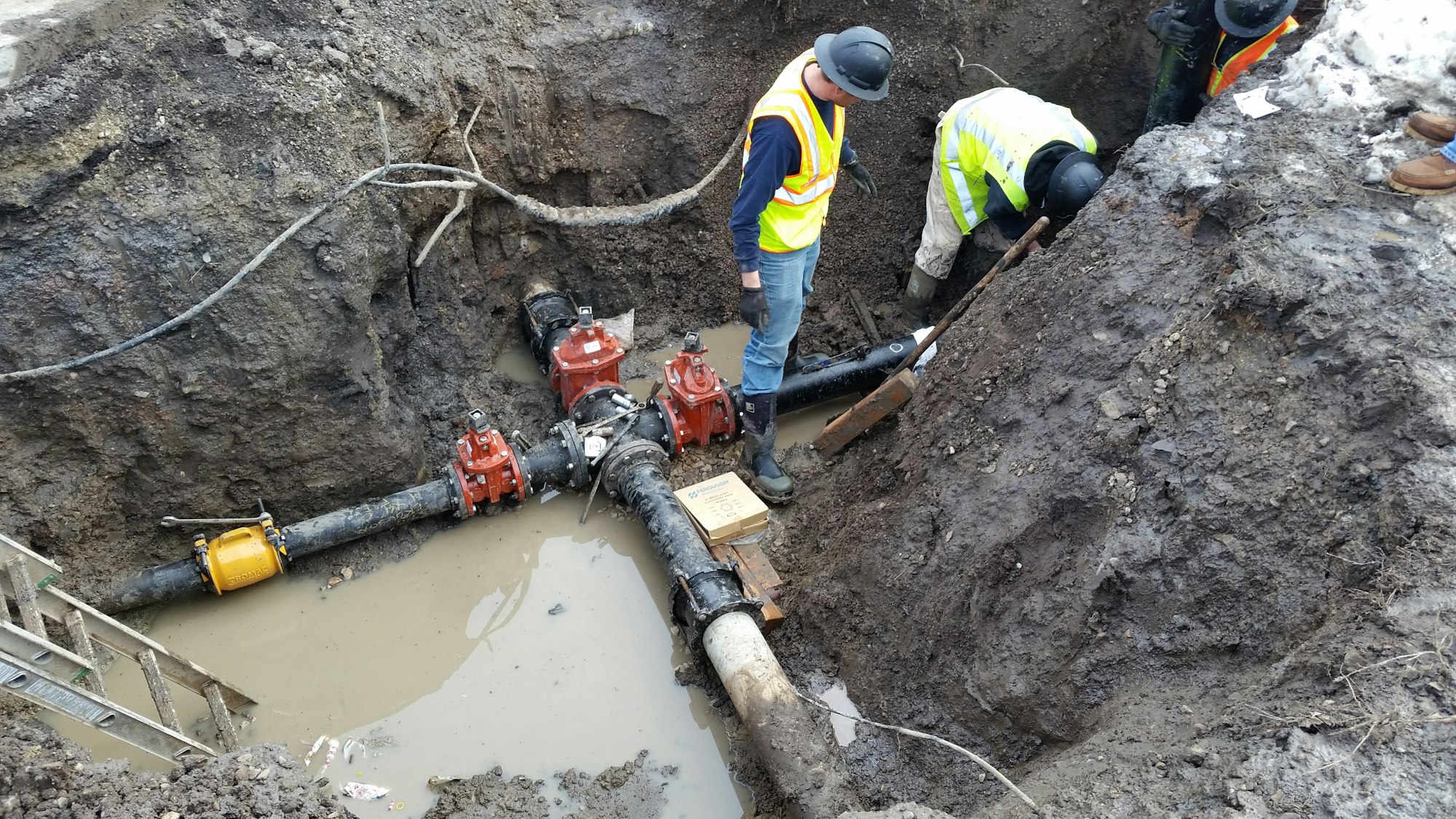 Workers in a trench working on underground pipes with visible water valves and muddy ground.