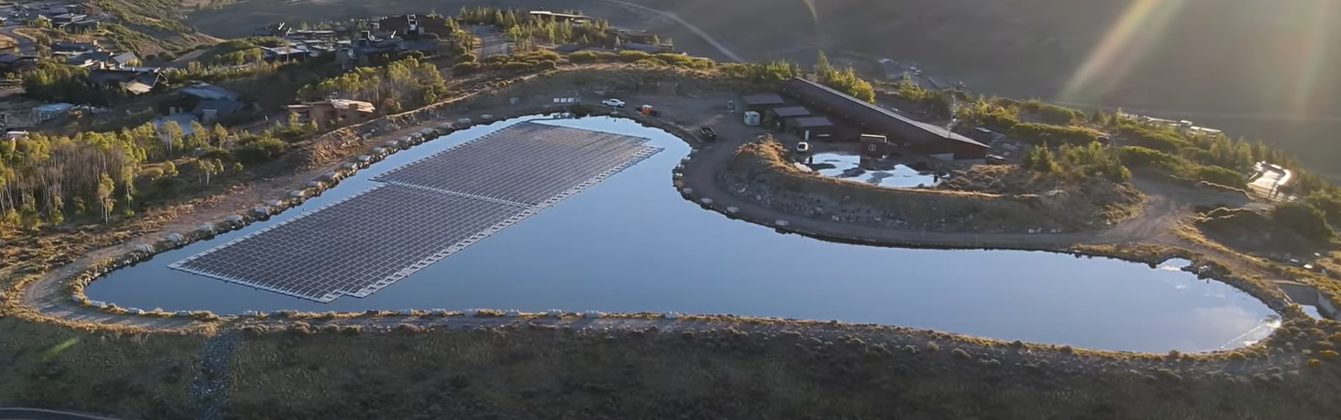 Aerial view of a water reservoir with solar panels, surrounded by wooded area and buildings, bathed in soft sunlight.
