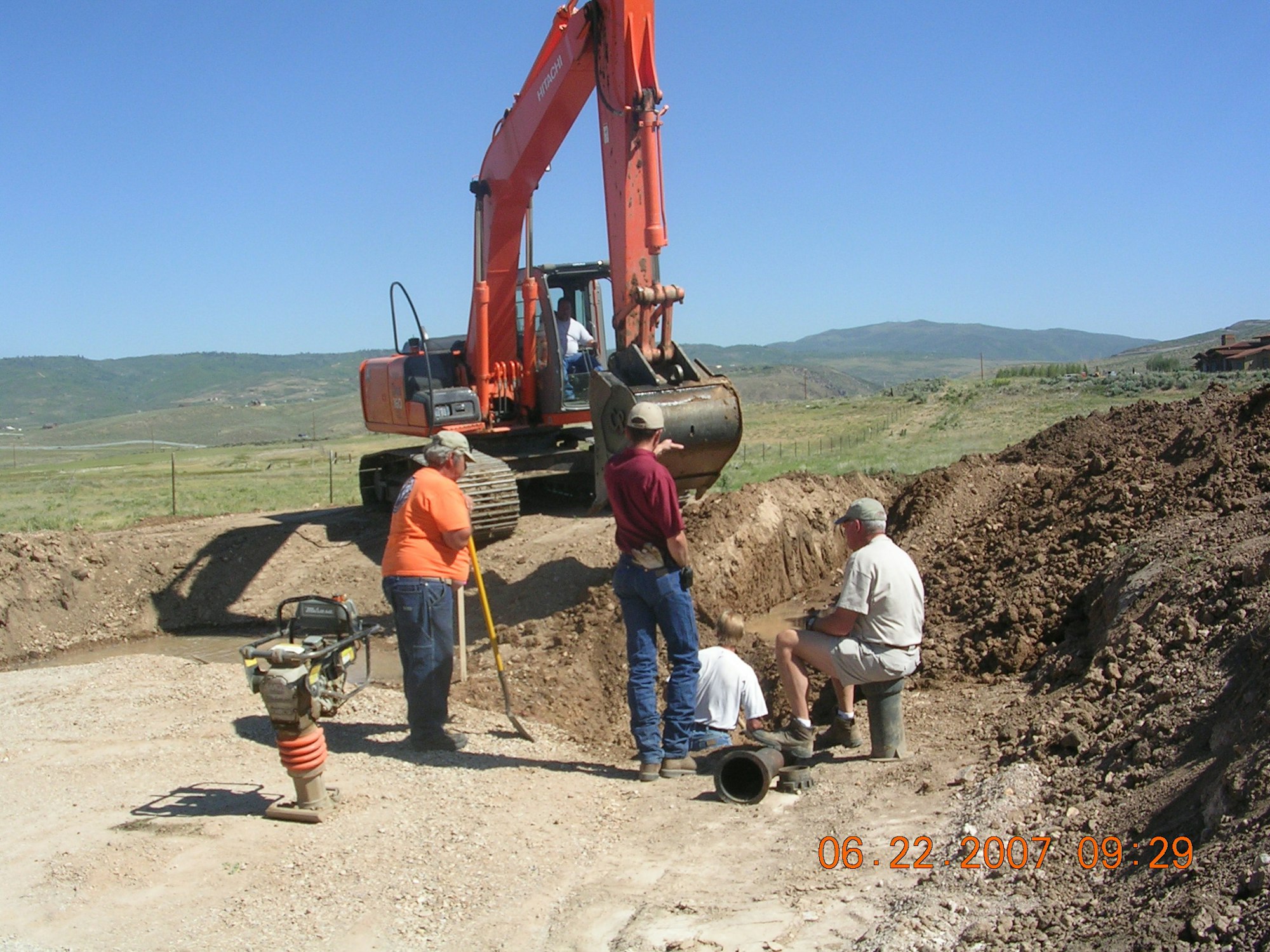 Construction site with a digger and workers discussing a pipe in a trench.