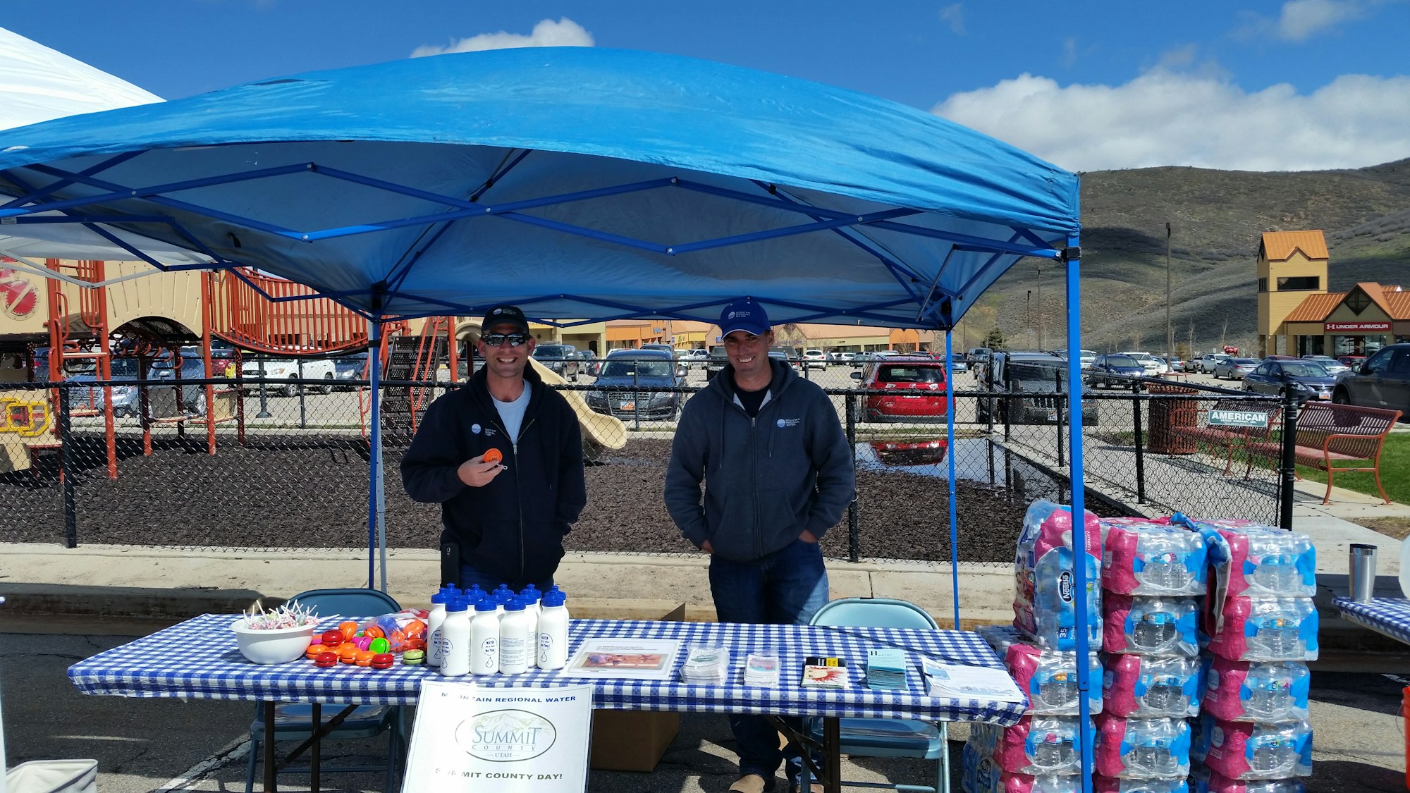 Outdoor booth with two people, water bottles, pamphlets, and a checkered tablecloth under a blue canopy.