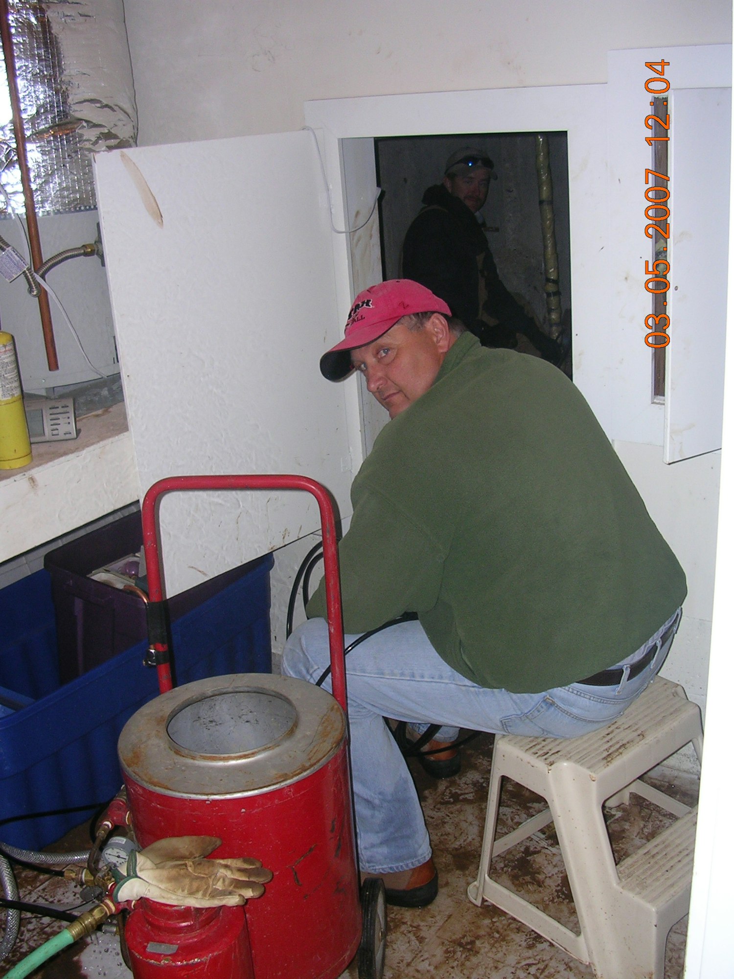 A man in a pink hat and green jacket sitting with equipment in a small room; another person is visible through a door.