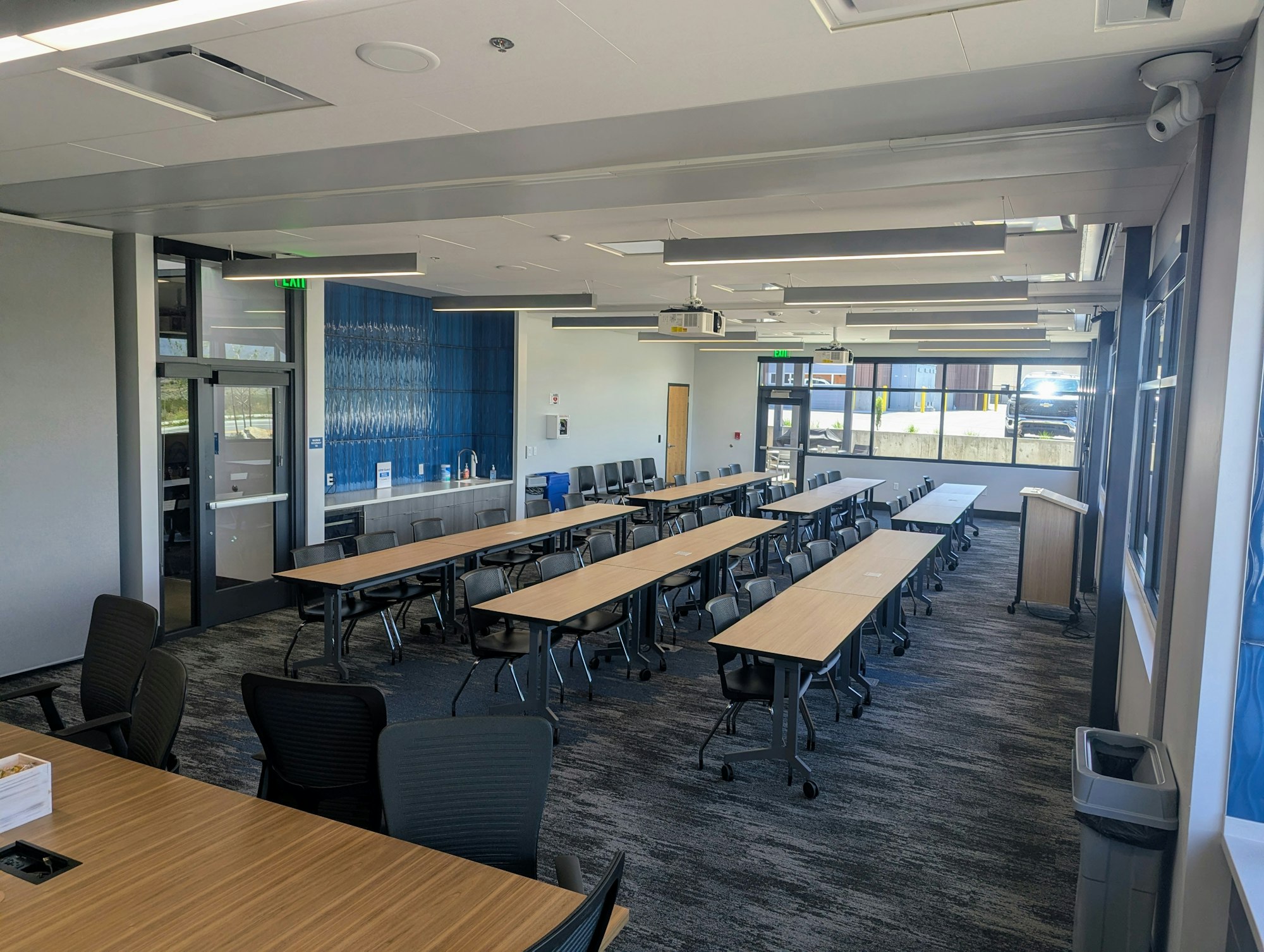 The image shows a modern classroom or meeting space with multiple tables, chairs, large windows, and a blue accent wall.