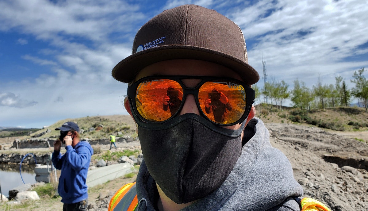 Person in hat and mask wearing reflective sunglasses, outdoors. Reflection shows person taking photo. Another person and landscape in background.