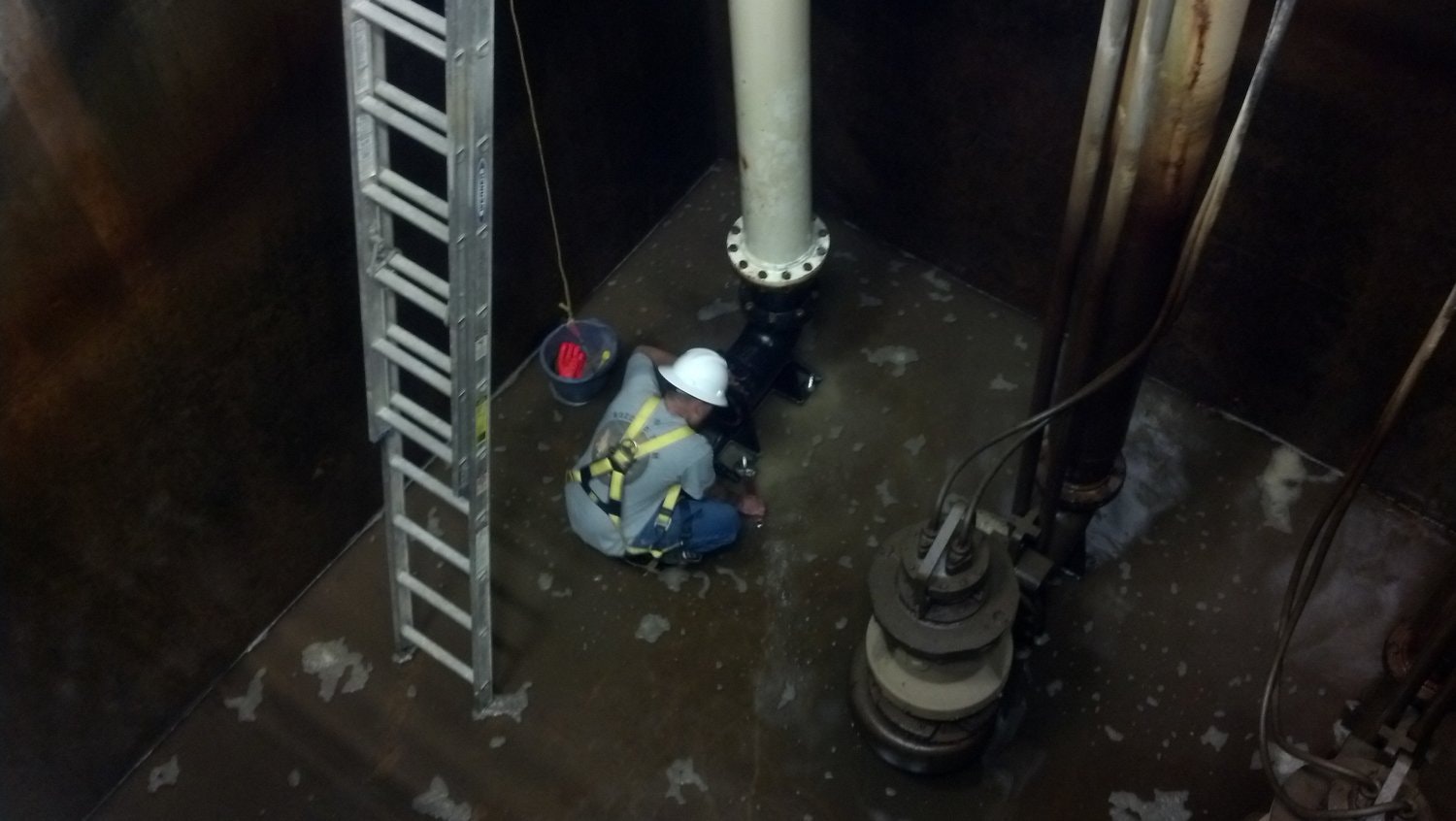 A worker in safety gear is repairing equipment in a dark, confined space, with a ladder and tools nearby.