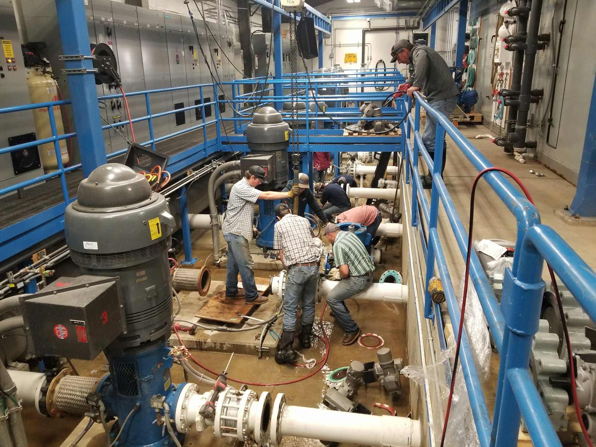 Workers are repairing or maintaining equipment in an industrial setting with large machinery and pipes.