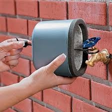 A person installs a faucet cover on an outdoor tap attached to a brick wall.