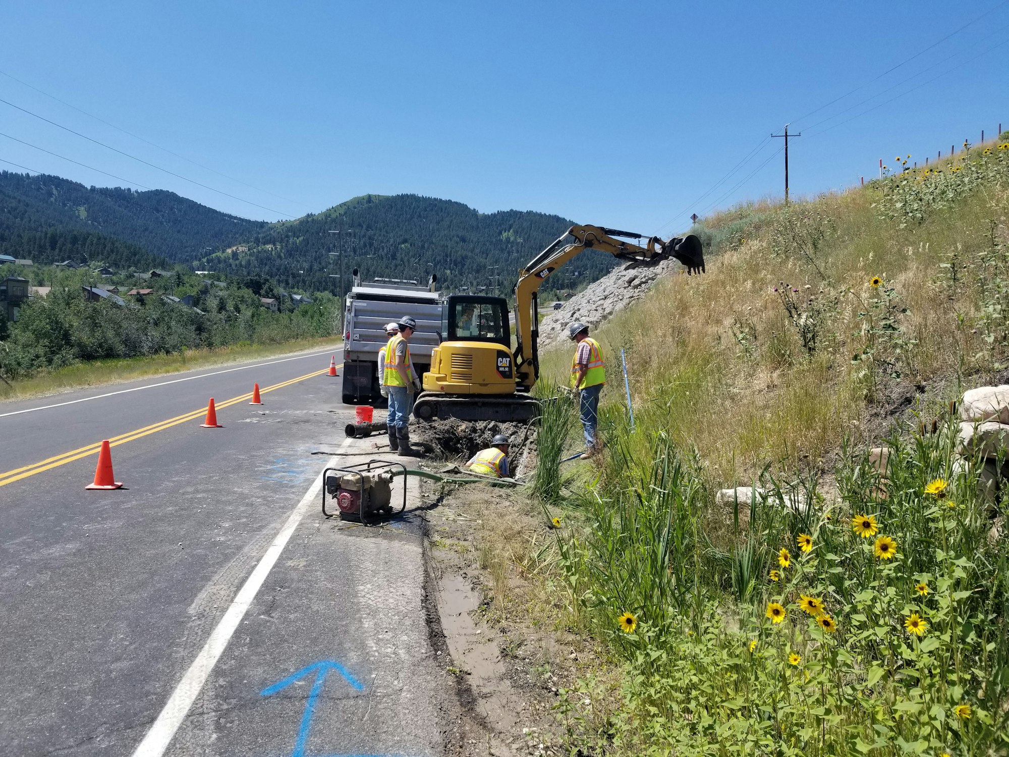 Road construction with workers, an excavator, and traffic cones along a rural road with mountains in the background.
