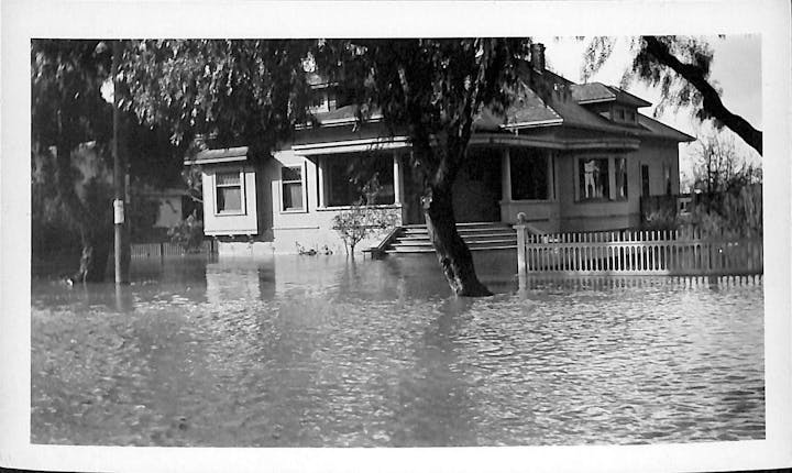A house partially submerged in water, indicating significant flooding, with nearby trees and a fence visible.