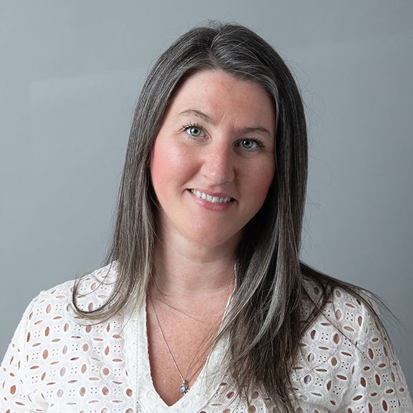A woman with long hair in a white patterned blouse, smiling against a gray background.