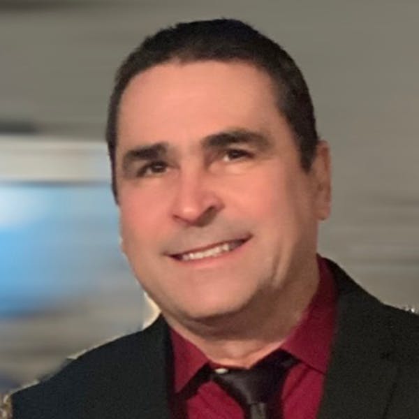 A person smiling, wearing a red shirt and dark jacket, is captured against a blurred background.