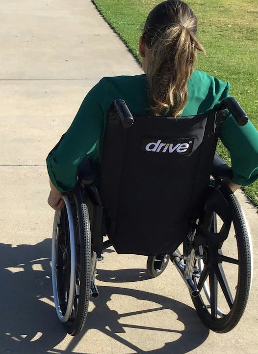 A person with long hair in a ponytail, wearing a green top, is using a "Drive" wheelchair on a paved path with grass beside it.