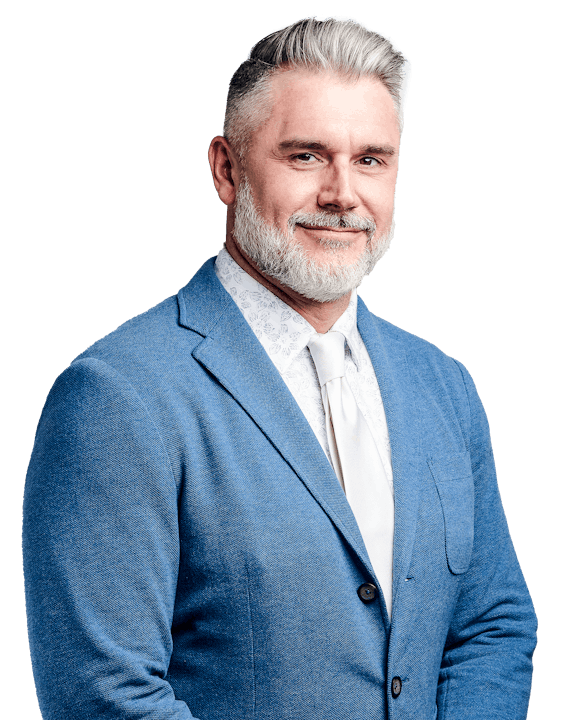 A person with gray hair and a beard, wearing a blue suit, white shirt, and tie, smiling against a black background.