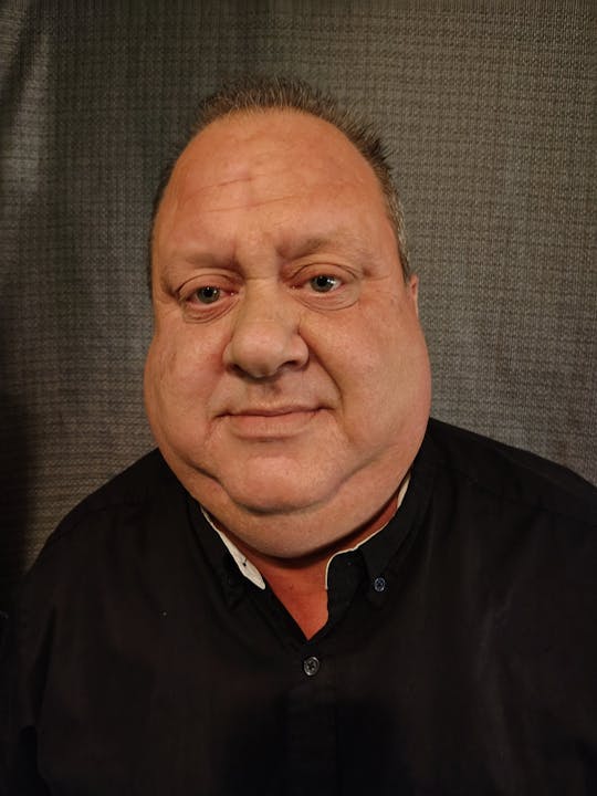 A man in a black shirt is looking directly at the camera against a textured background.
