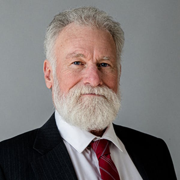 An older man with a white beard, wearing a suit and red tie, against a gray background.