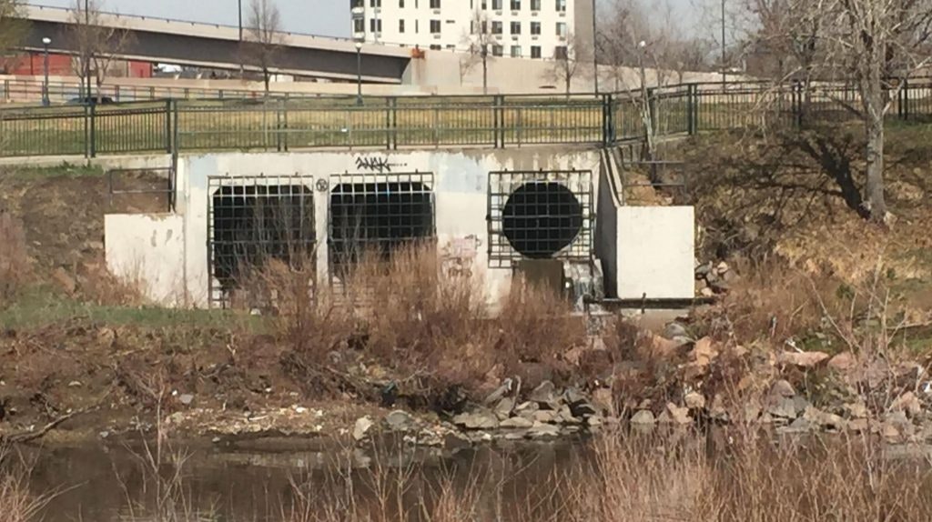 Twin culverts with barred entrances beside a body of water, graffiti on the facade, with a backdrop of grass and a fence.