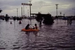 Flooded street with people in a dinghy and others wading through water, street signs and vehicles partially submerged.