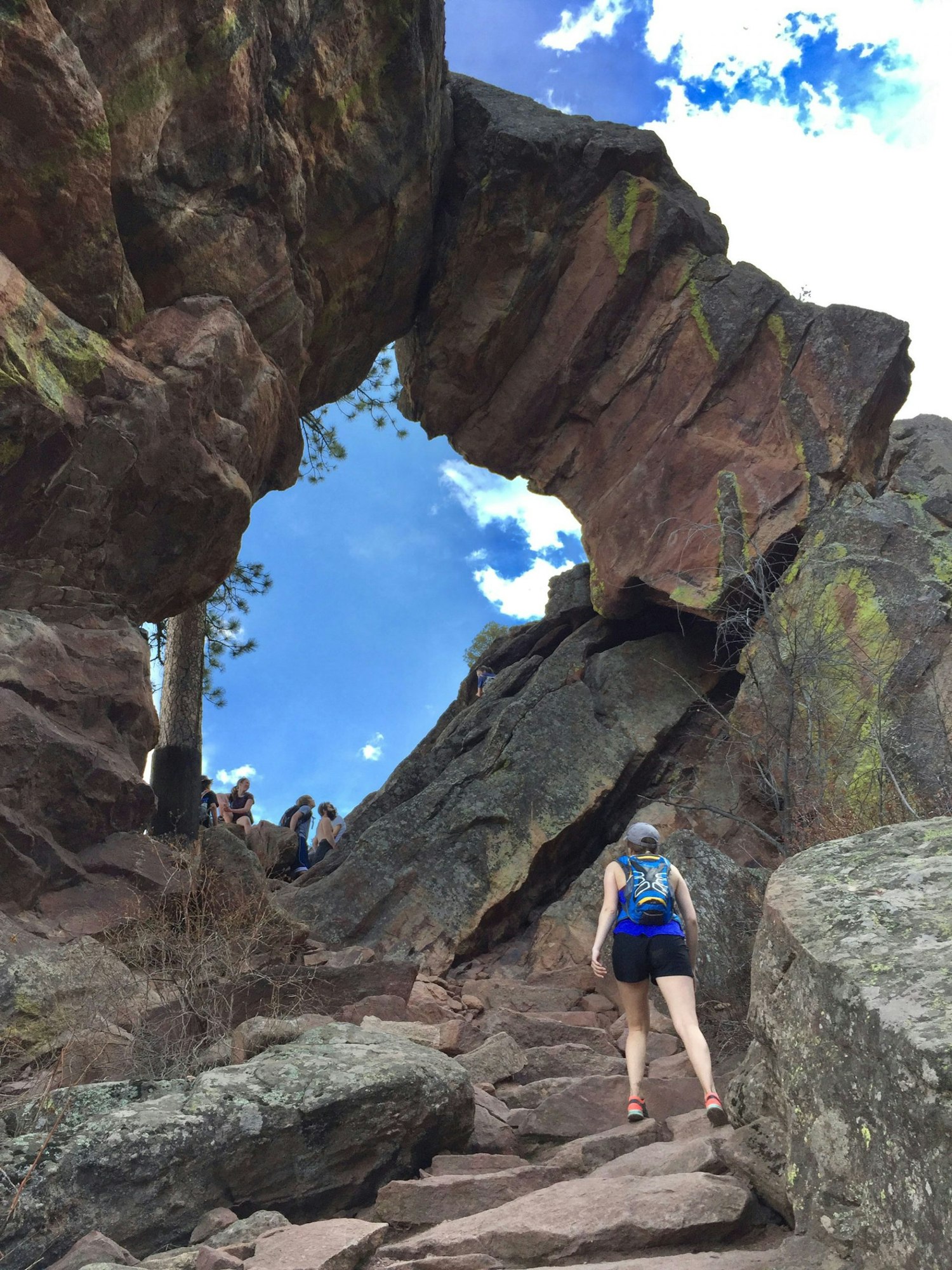 A hiker approaches a natural rock arch, with others climbing above on a rocky path under a blue sky.