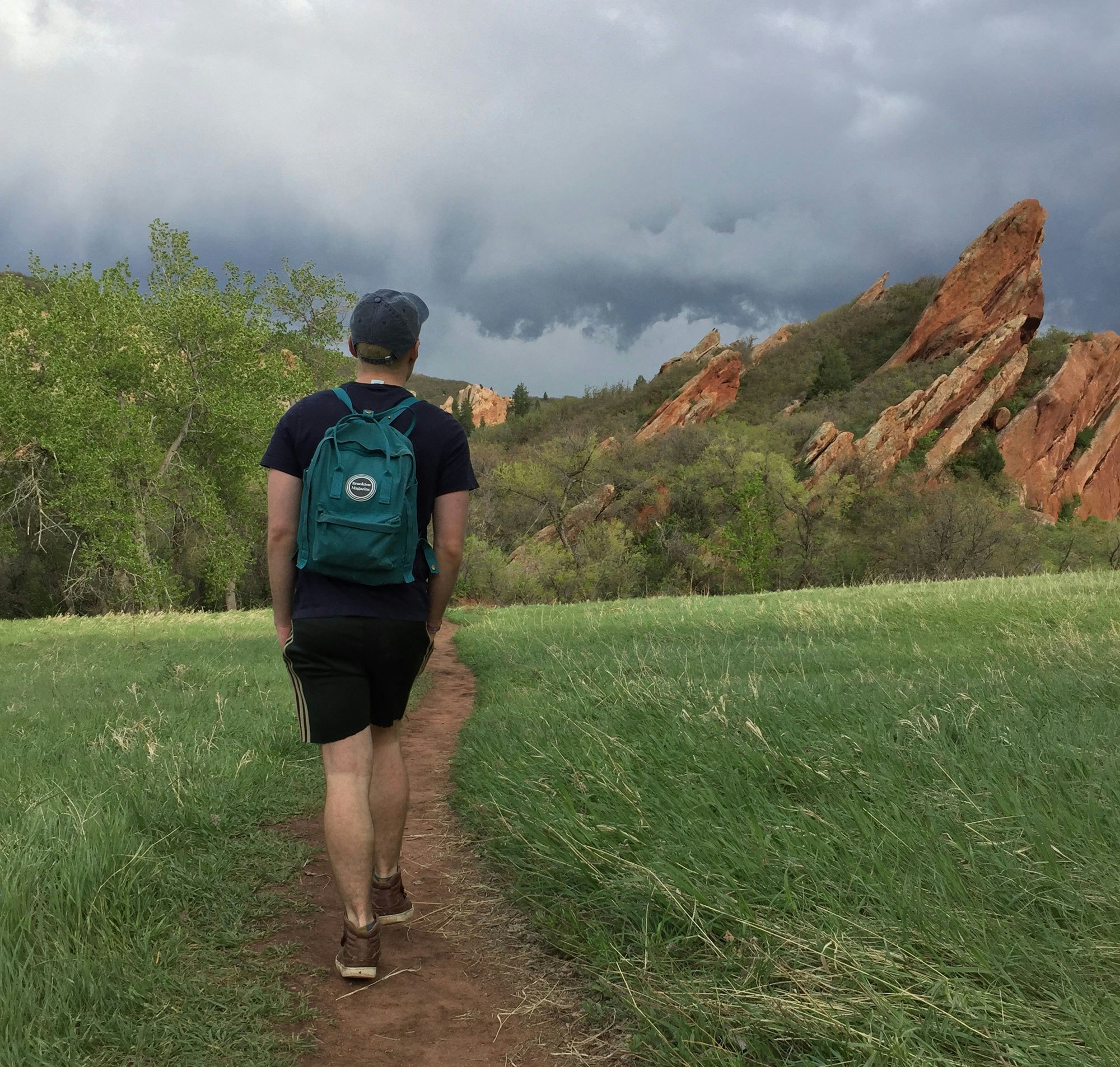 Person hiking a trail towards rocky formations under a stormy sky.