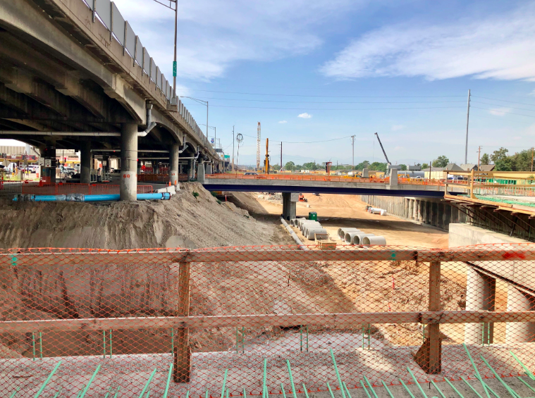 A construction site with excavated earth, concrete pillars, safety fences, and a crane under a skyway bridge.