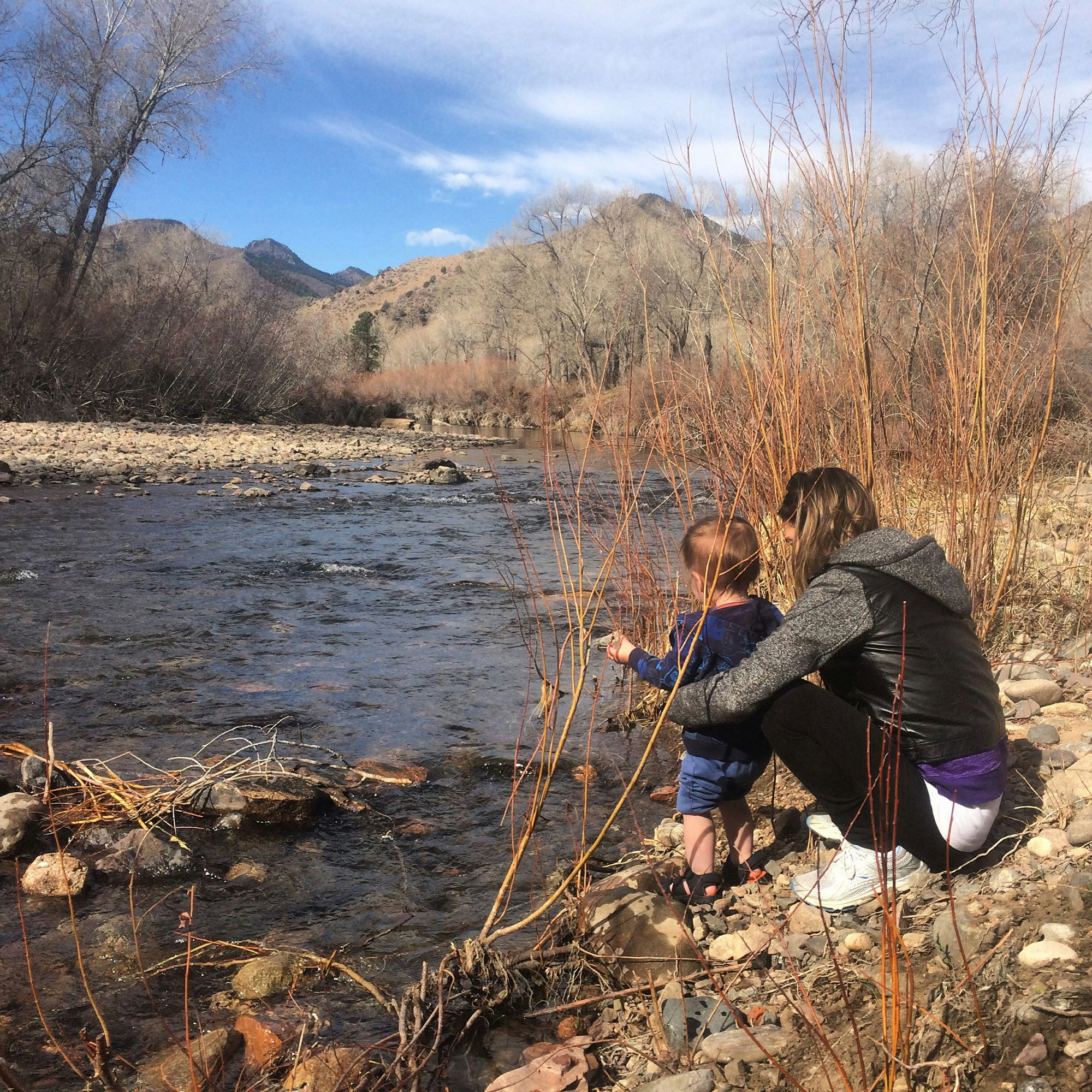 A child and an adult by a river with mountains in the background.