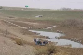 Rural landscape, people by a small water body, underpass, green sign in distance, overcast sky.