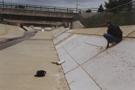 A man sitting by a dry canal with a bridge overhead and debris on the ground.