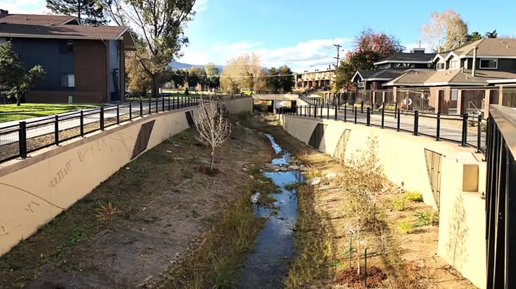 A dry urban canal with a small water stream, surrounded by fences, trees, and residential buildings under a clear sky.