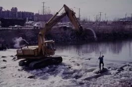 An excavator near water with a person standing close by, with urban background.
