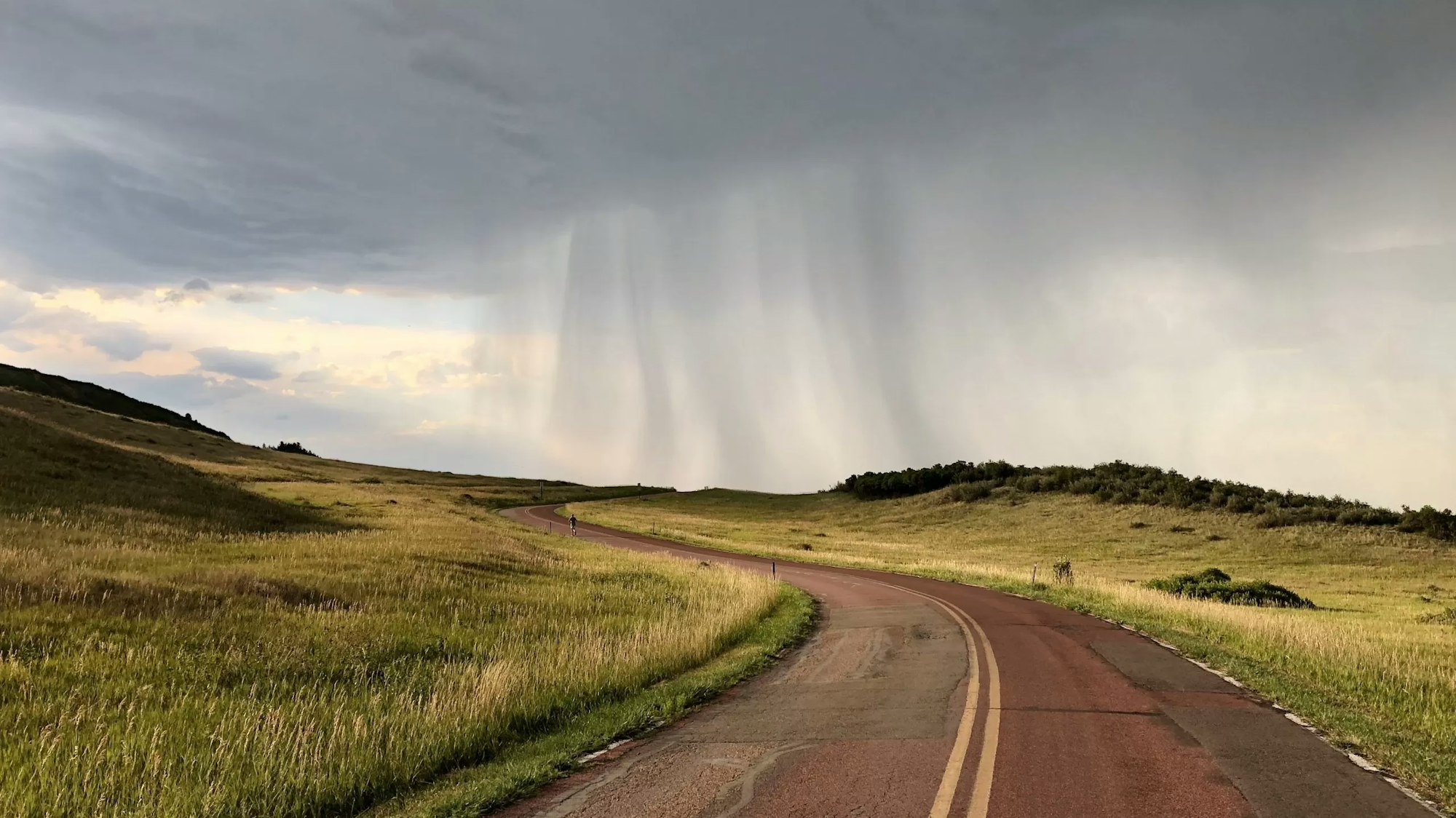 A winding road through a grassy landscape with a dramatic rain shower in the background under a gloomy sky.