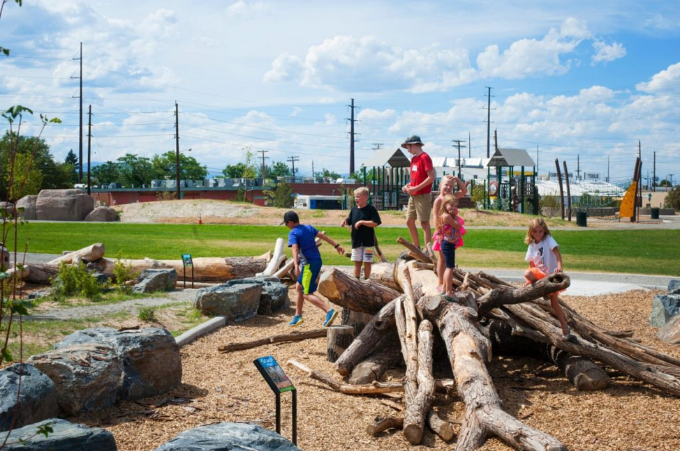 Children and adults at a playground with logs and rocks on a sunny day.