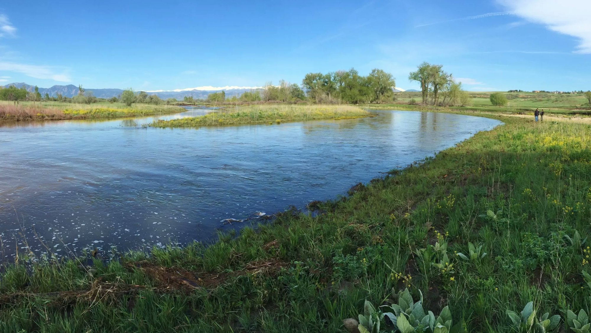 A serene river landscape with greenery and distant mountains, under a clear blue sky, with a couple walking by the water.