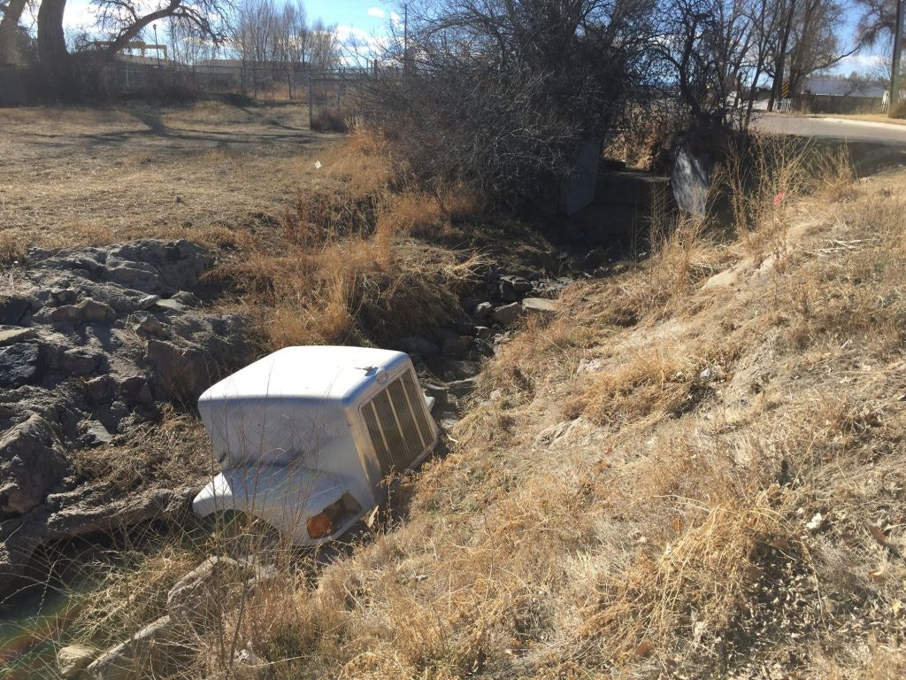 A discarded washing machine in a dry creek bed with rocks and sparse grass.