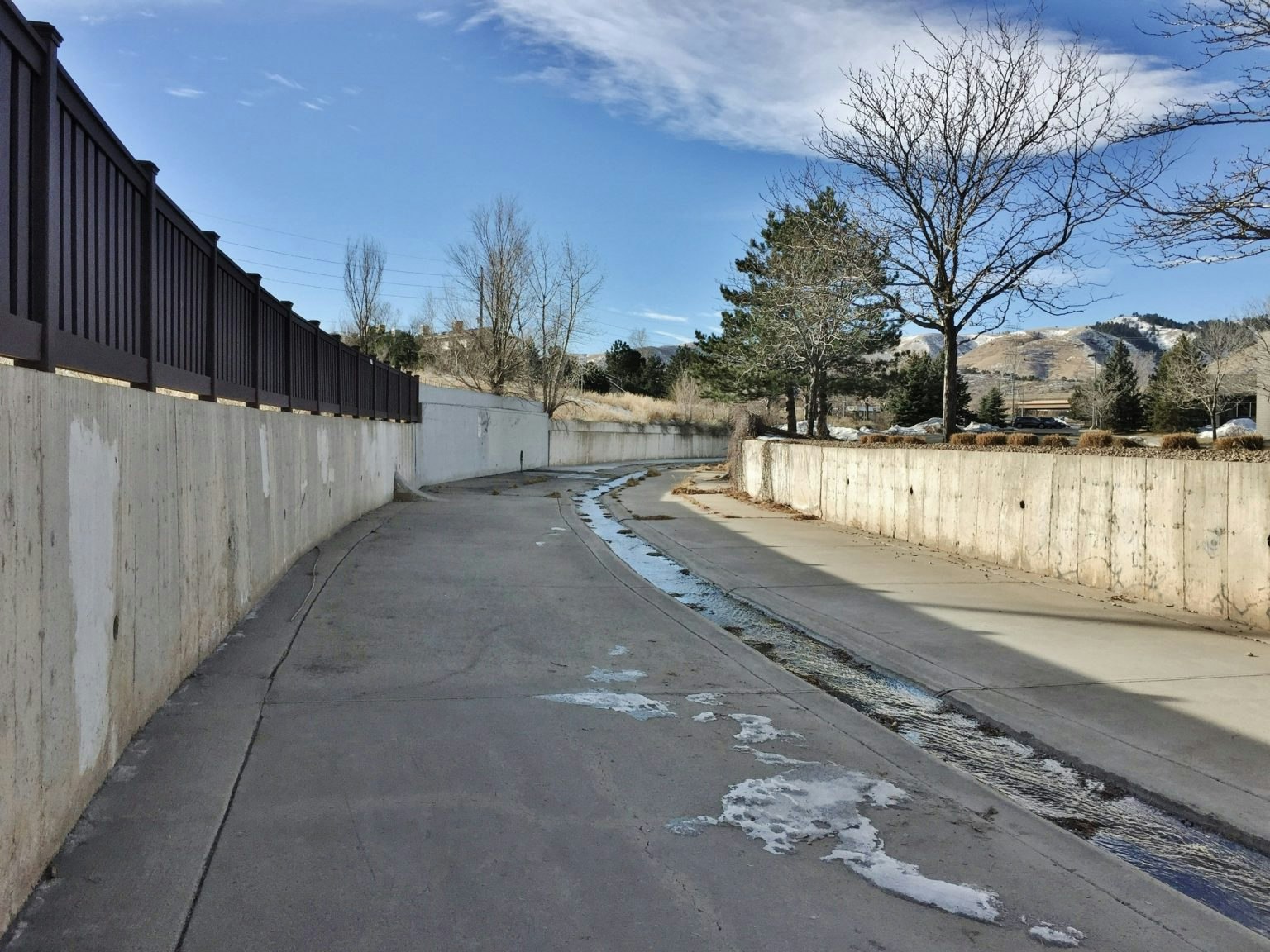 A walkway with a metal fence and concrete barrier, bare trees, snow patches, and distant hills.