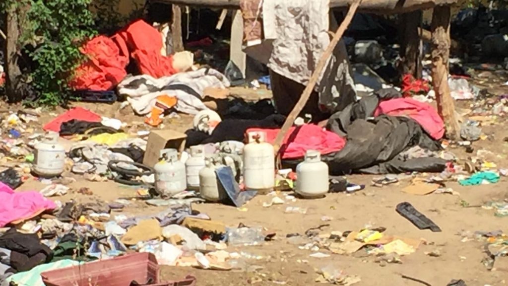 A cluttered area with trash, clothing, and containers scattered around an informal shelter structure.