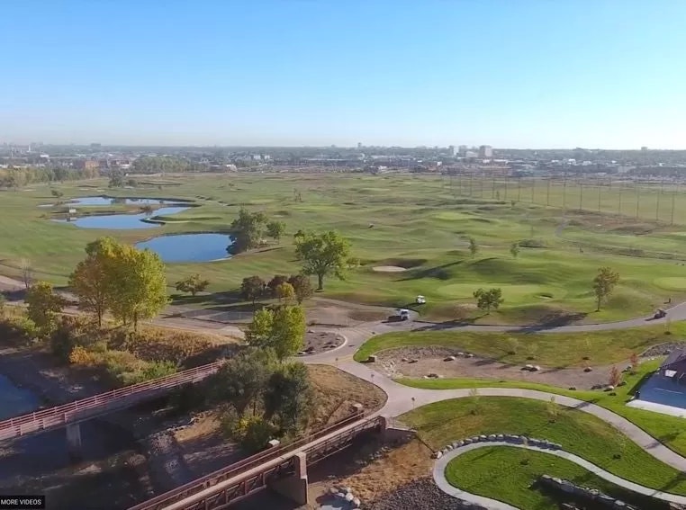 Aerial view of a golf course with ponds, greenery, and a city skyline in the background.