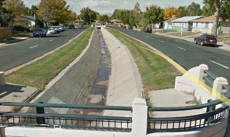 Suburban street with a dividing drainage ditch, cars parked, and trees lining the sidewalks.